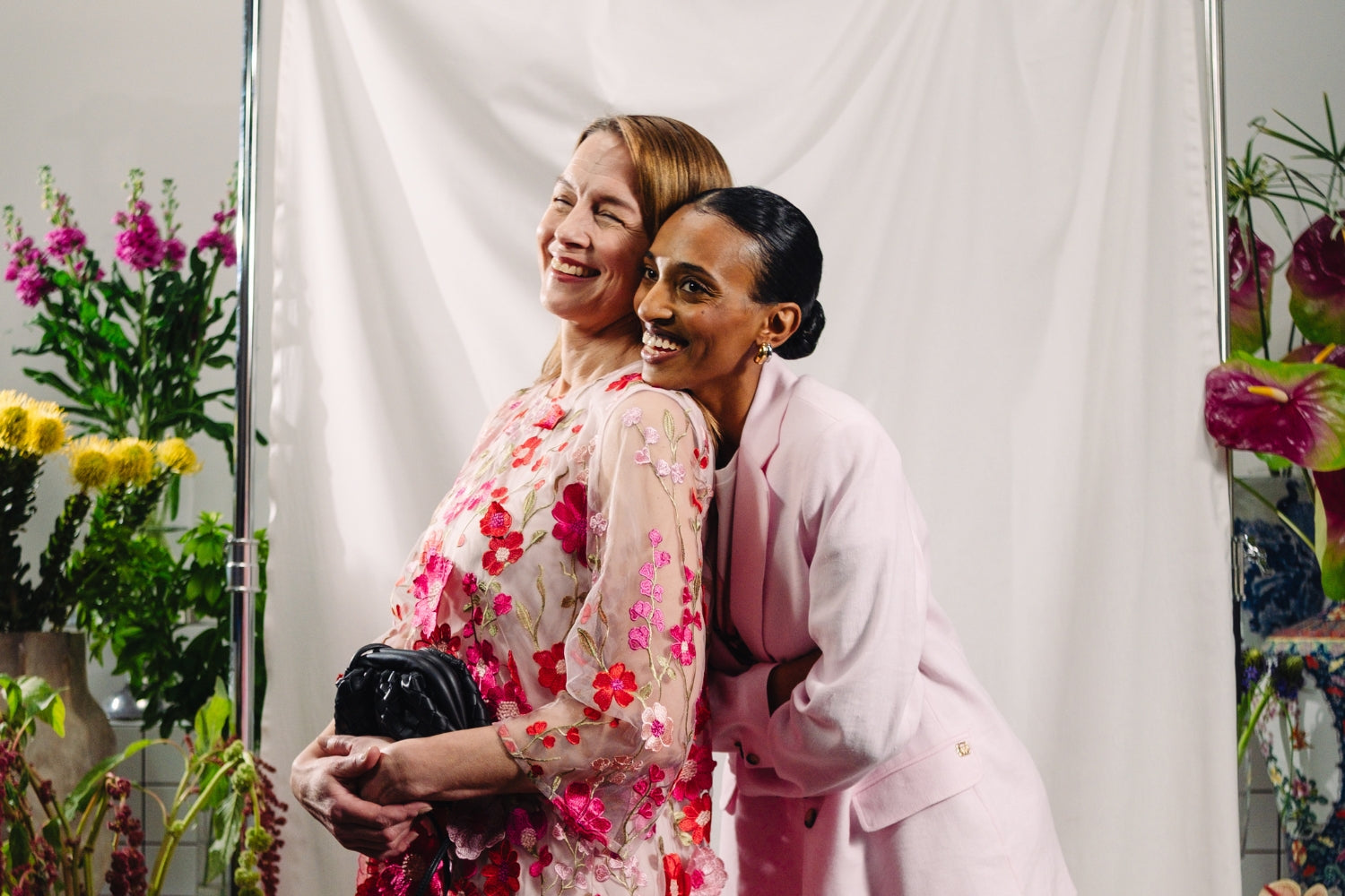 Kaksi iloista naista taustallaan vaalea kangas ja kukkia. Toisella yllään kukkamekko ja toisella jakkupuku.