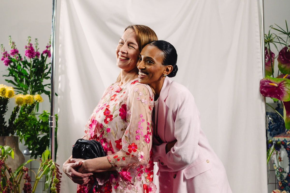 Kaksi iloista naista taustallaan vaalea kangas ja kukkia. Toisella yllään kukkamekko ja toisella jakkupuku.