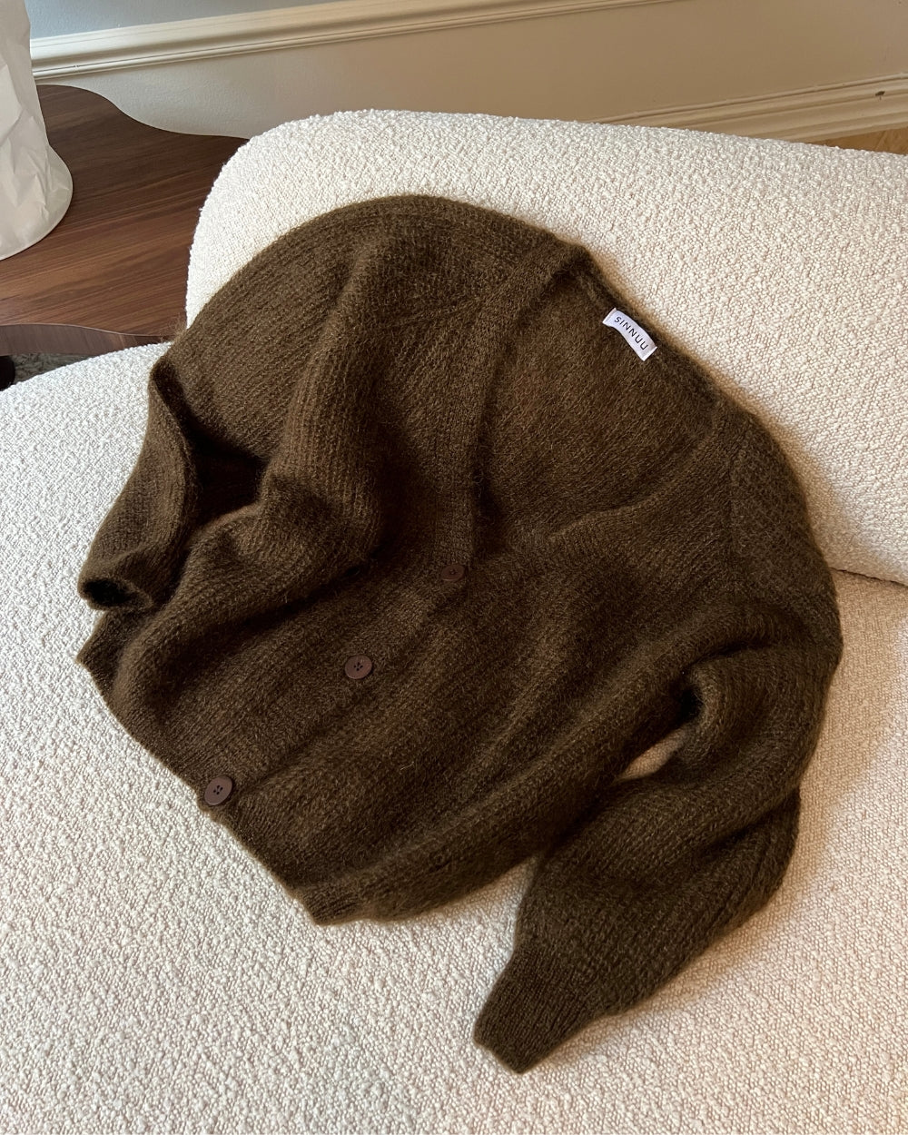 Tummanruskea, pörröinen neuletakki vaalealla sohvalla.