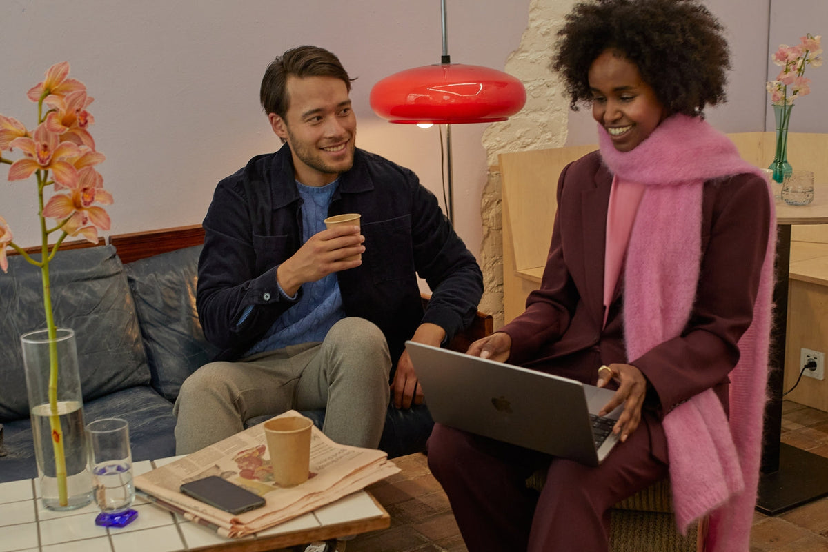 Mies ja nainen istuvat kahvilassa. Naisella on sylissään kannettava tietokone ja hän on pukeutunut viininpunaiseen jakkupukuun sekä vaaleanpunaiseen huiviin. Miehellä on kädessään kahvikuppi ja hän on pukeutunut chino-housuihin ja paitatakkiin.