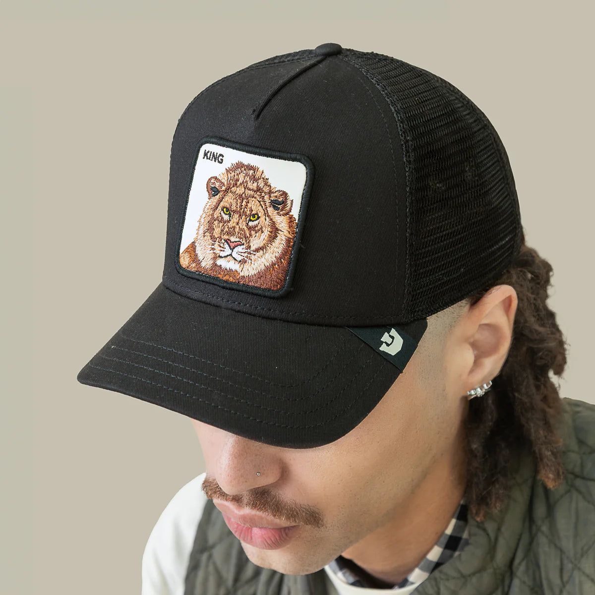 Goorin Bros lippis Lion King Trucker, musta, jossa musta verkkotaustainen takaosa, kiinteä lippa ja edessä suurehko, keltaisin ja ruskein sävyin brodeerattu leijonakuvio, laadukas materiaali ja suoraviivainen muotoilu.